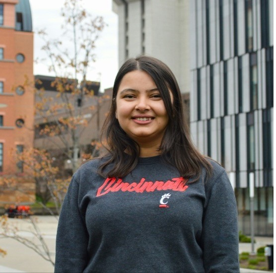 A smiling photo of Garima Mandavya with dark hair, wearing a University of Cincinnati long-sleeve shirt, with buildings and trees in the background.