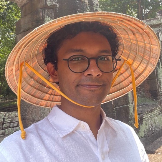 A smiling photo of Amartya Mitra wearing a hat, glasses, and a white collared shirt with trees in the background.