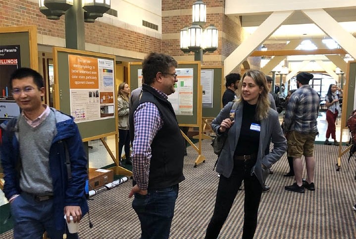 Conference attendees of Sensorium 2019 mingle in a large hallway in front of hanging lights and scientific poster boards.