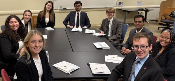 Students wearing business professional attire sit around conference room table, smiling and looking toward camera.