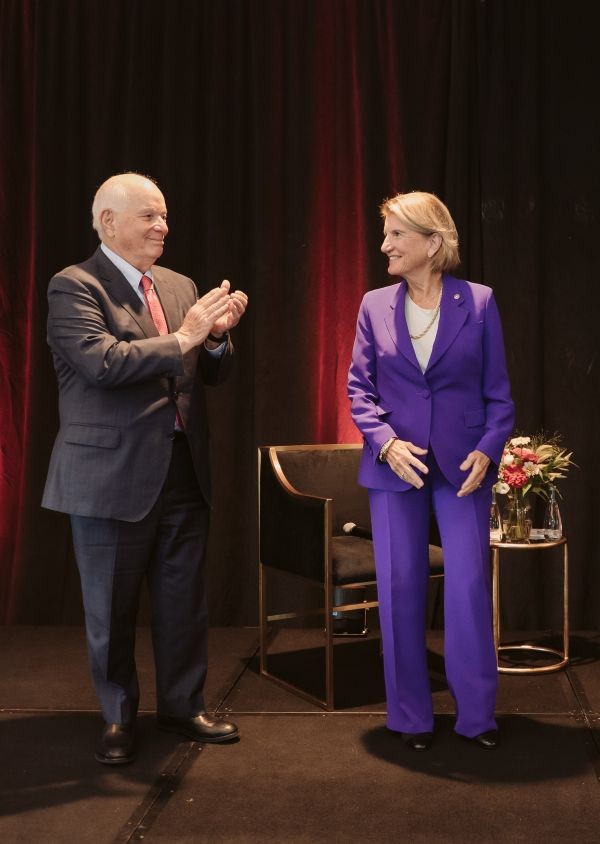 A man and woman stand on a stage with an elegant fabric backdrop. The man looks toward the woman and smiles; he claps his hands in applause.  Both wear polished business suits.