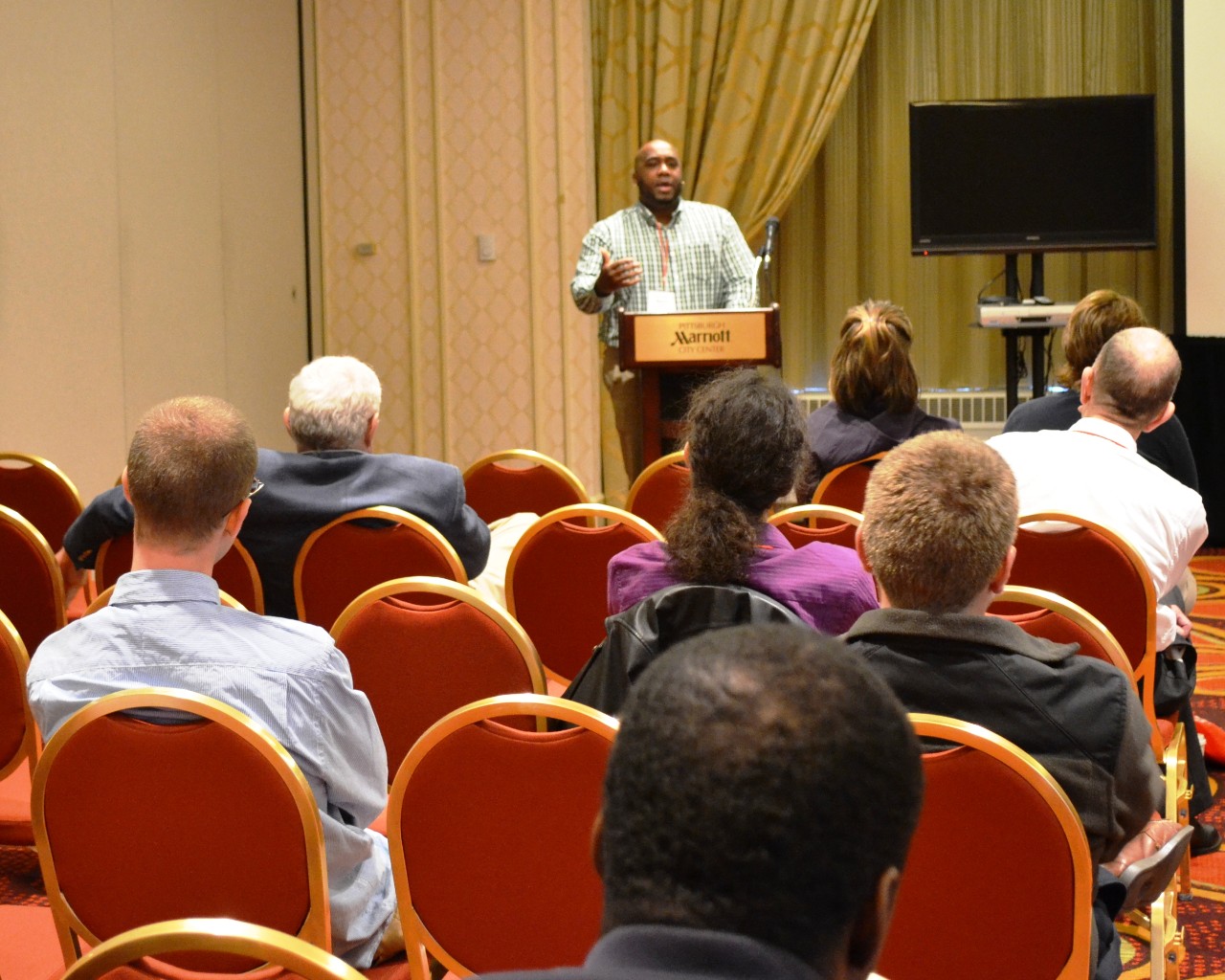 Man standing at podium speaking to a room full of people sitting in chairs