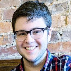 A smiling color photo of Michael Meece wearing a flannel shirt in front of a brick background.