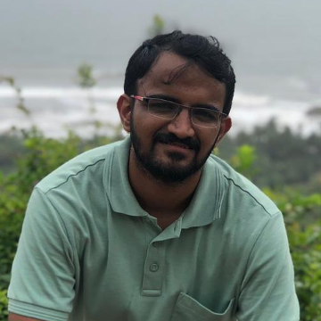 A smilingn photo of Abhinav Madabhushi with dark hair, wearing glasses and a green collared shirt, with greren plants and an ocean in the background.