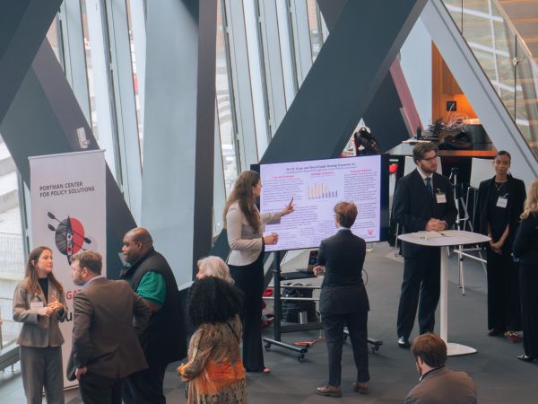 People stand in small groups in a building with dramatic beams and large windows. At the focus of each group an undergraduate student speaks to the people listening.
