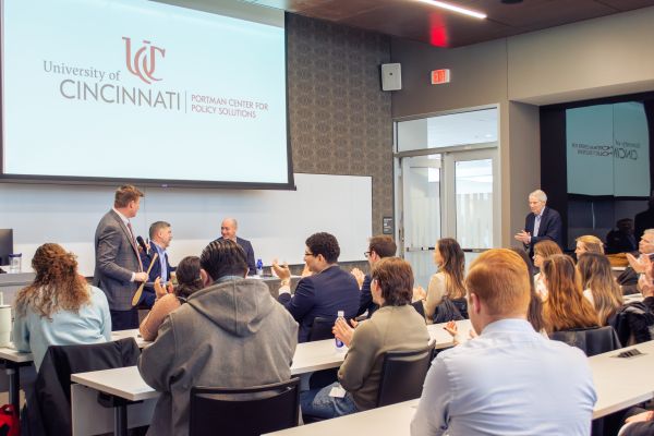 An audience sits facing the front of the row. Four men are in the front of the room; one stands and speaks into a microphone.
