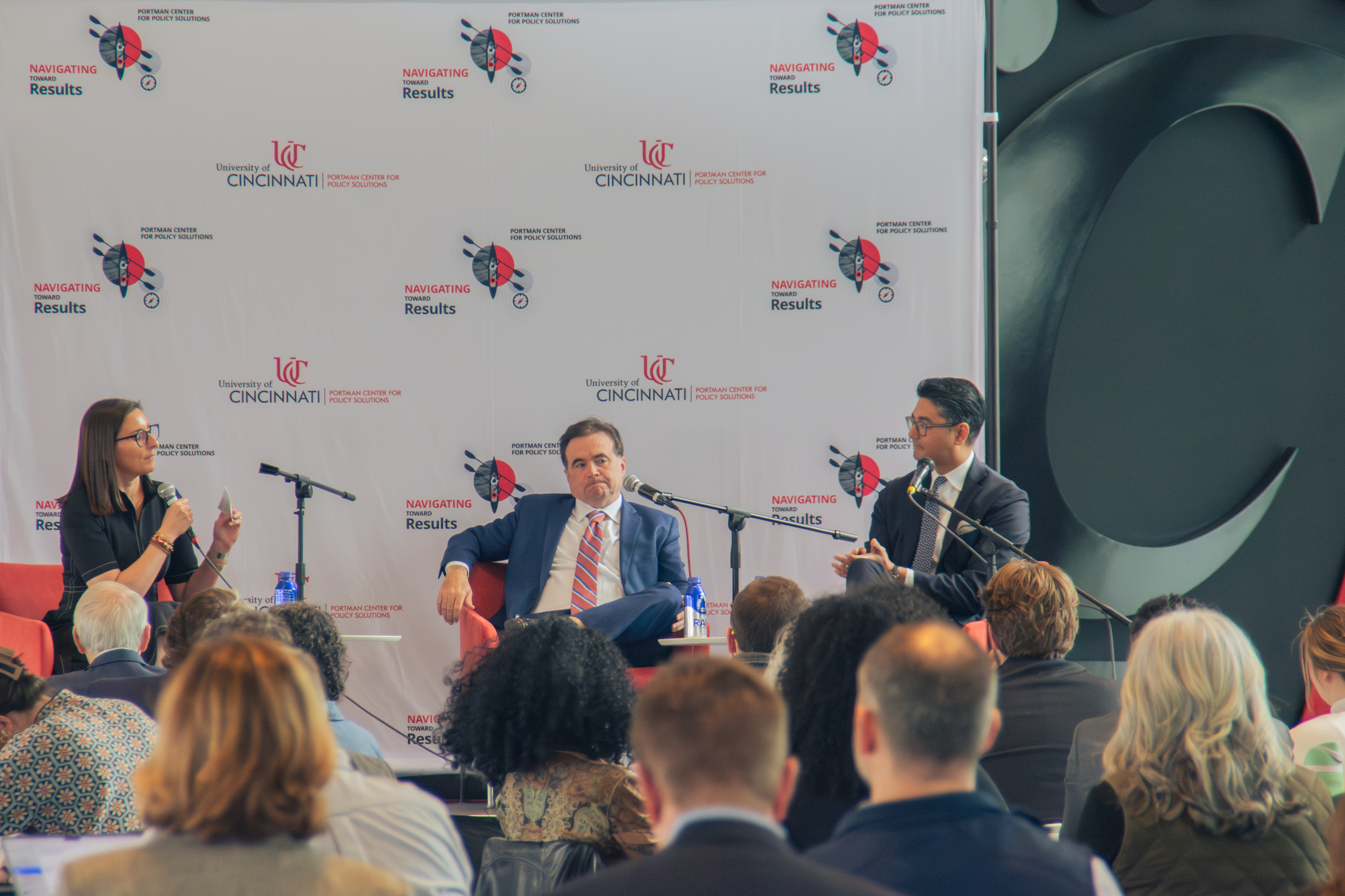 Three panelists in business attire engage in a discussion on stage at a University of Cincinnati event, with attentive audience members seated in front.