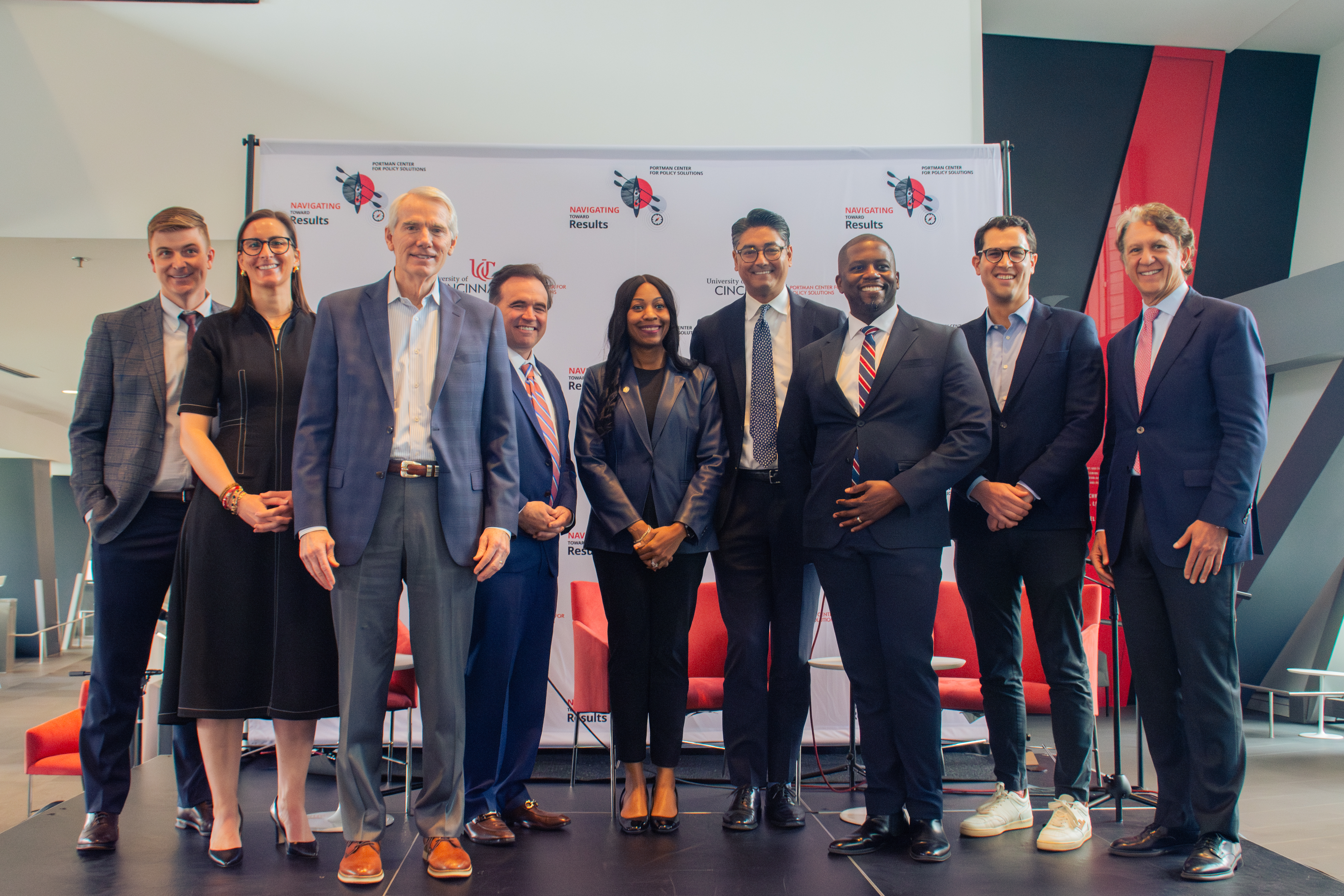A group of nine well-dressed individuals smiling and posing in front of a branded backdrop. The setting appears professional and upbeat.