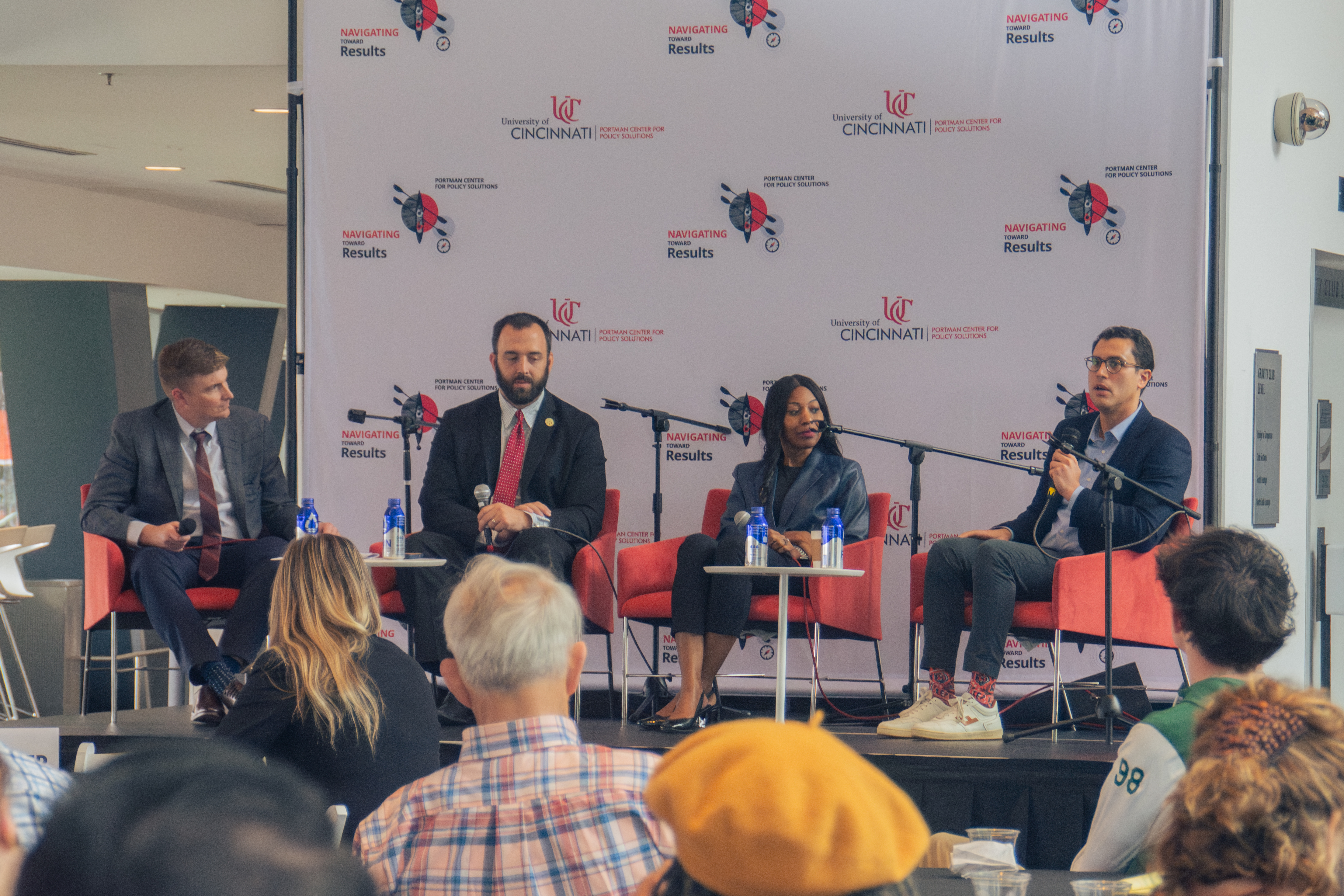 Four panelists, three men and a woman, sit on a stage with a University of Cincinnati backdrop. They are engaged in a discussion with an audience.