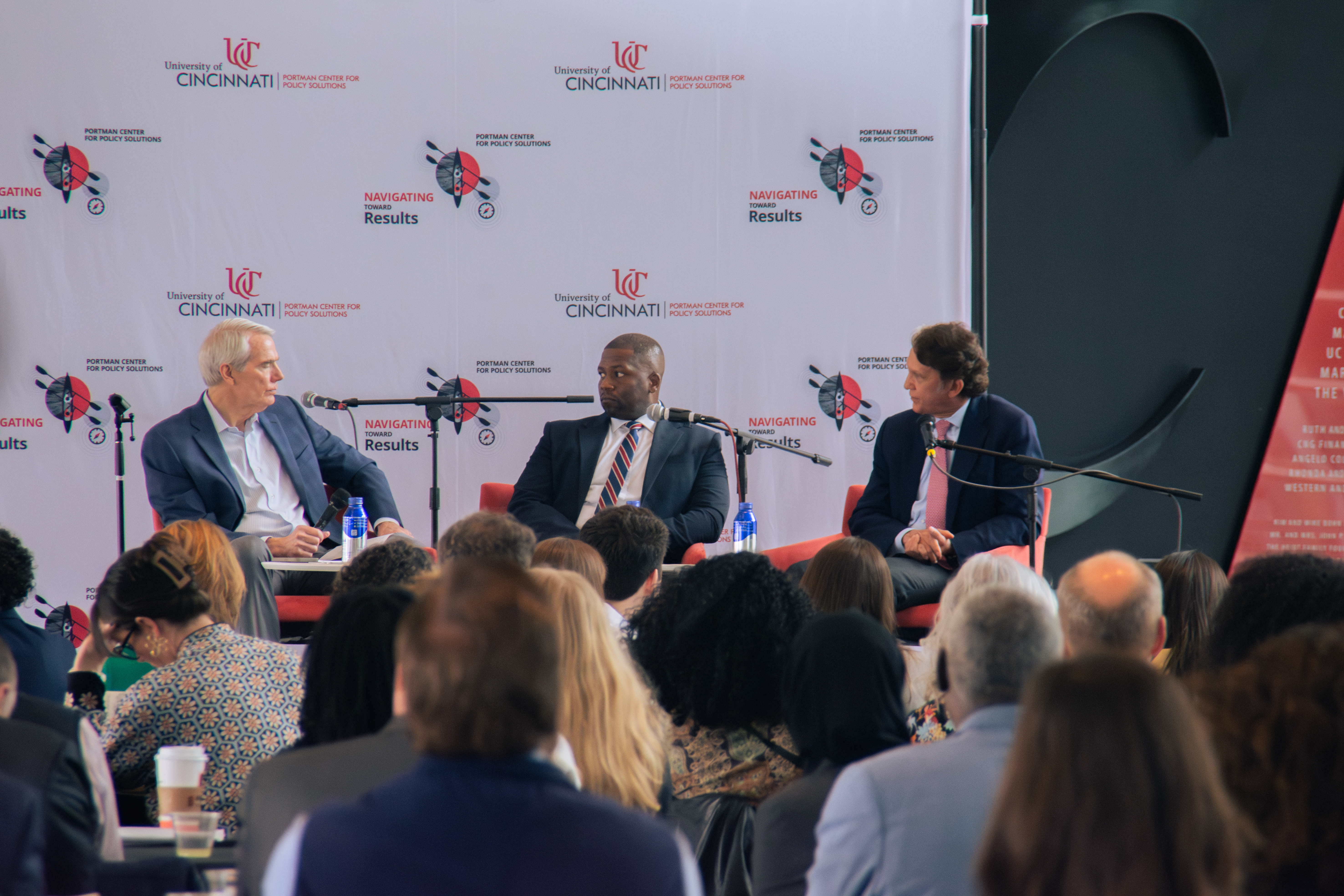 Three men in suits sit on stage at a University of Cincinnati event, discussing policy. Audience members listen attentively. Tone is professional.