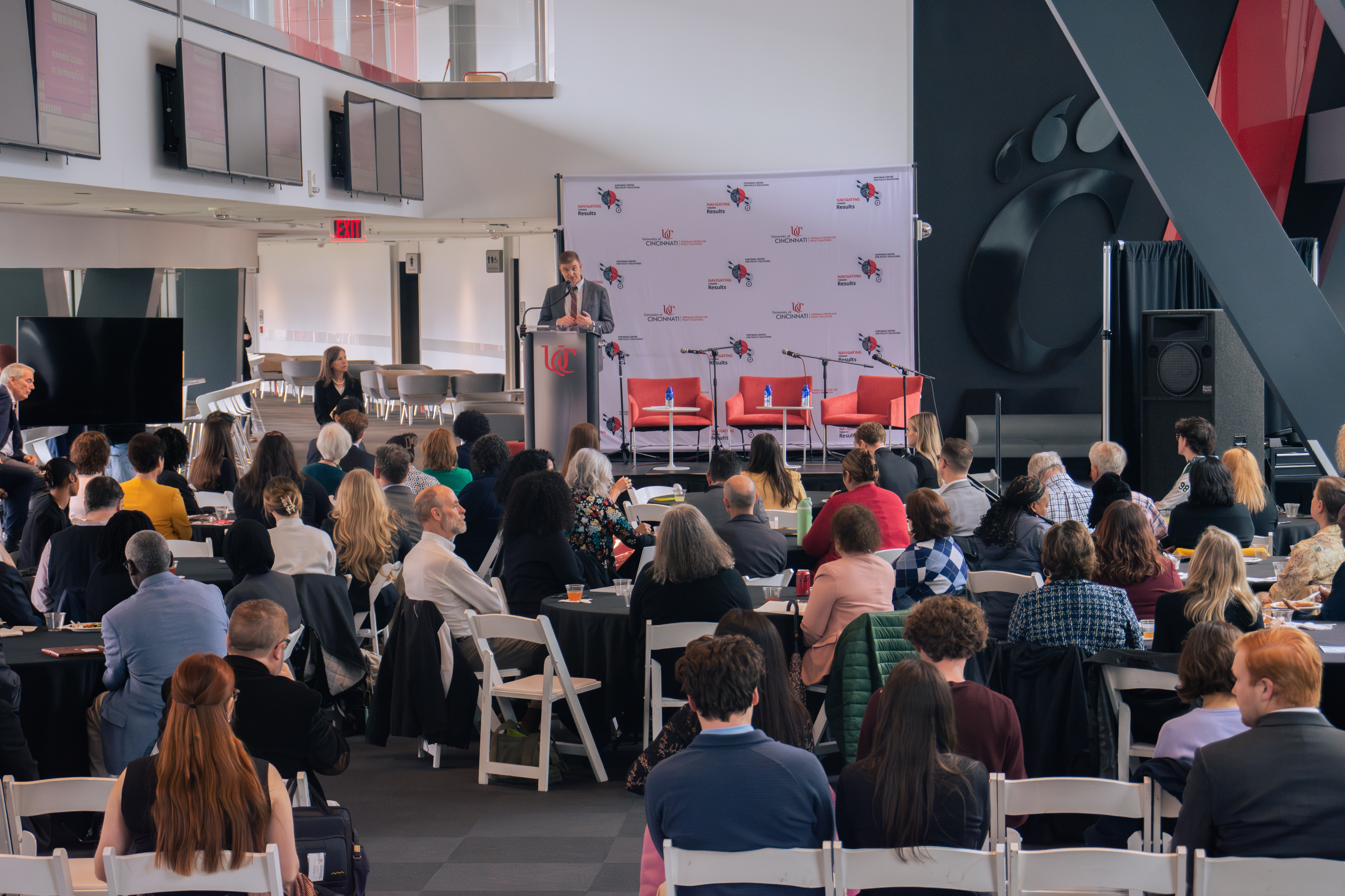 A speaker at a podium addresses a seated audience in a modern conference room. The setting is formal, with attentive attendees and a large backdrop.