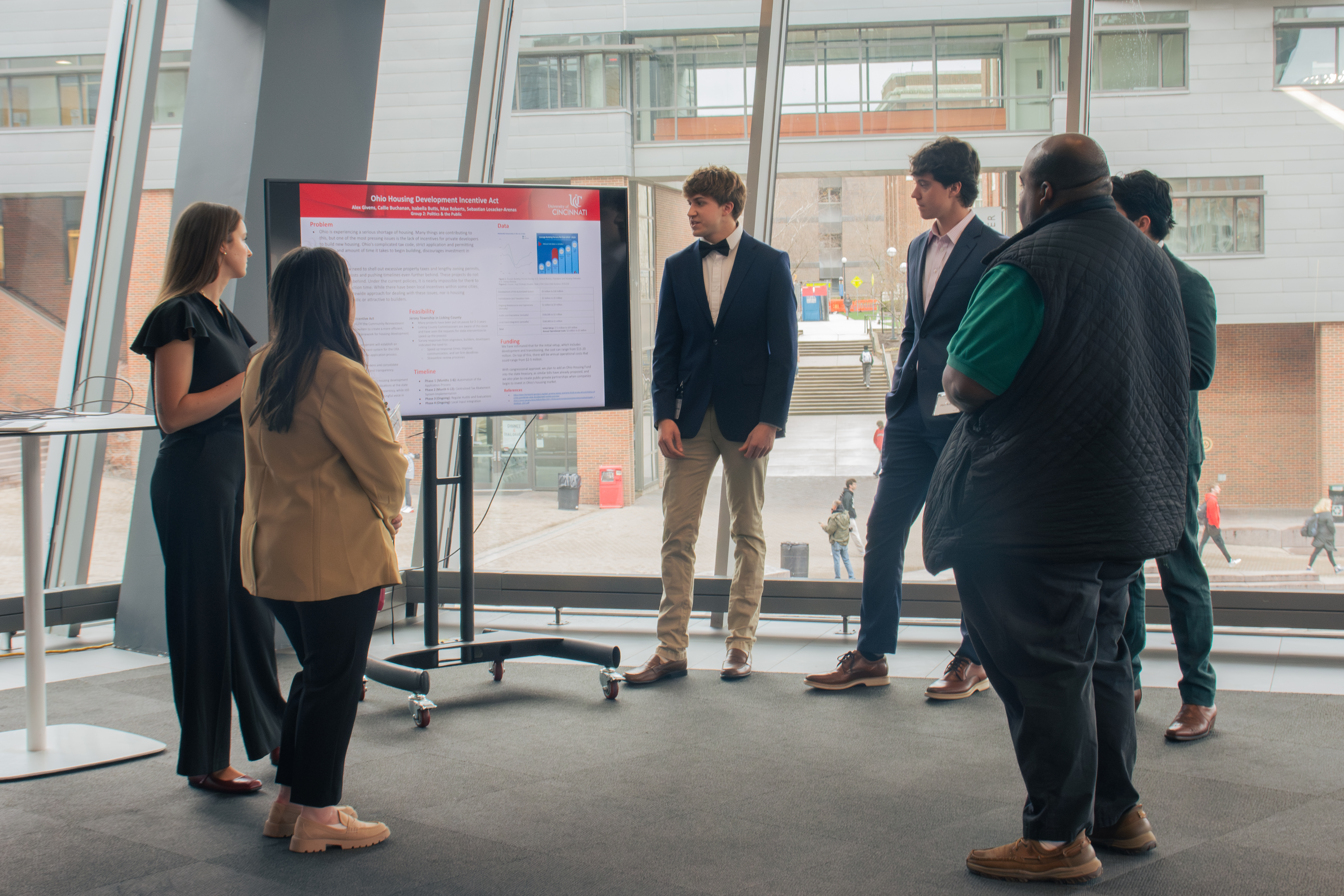 A group of people standing around student presenters listening to a presentation displayed on a screen. 