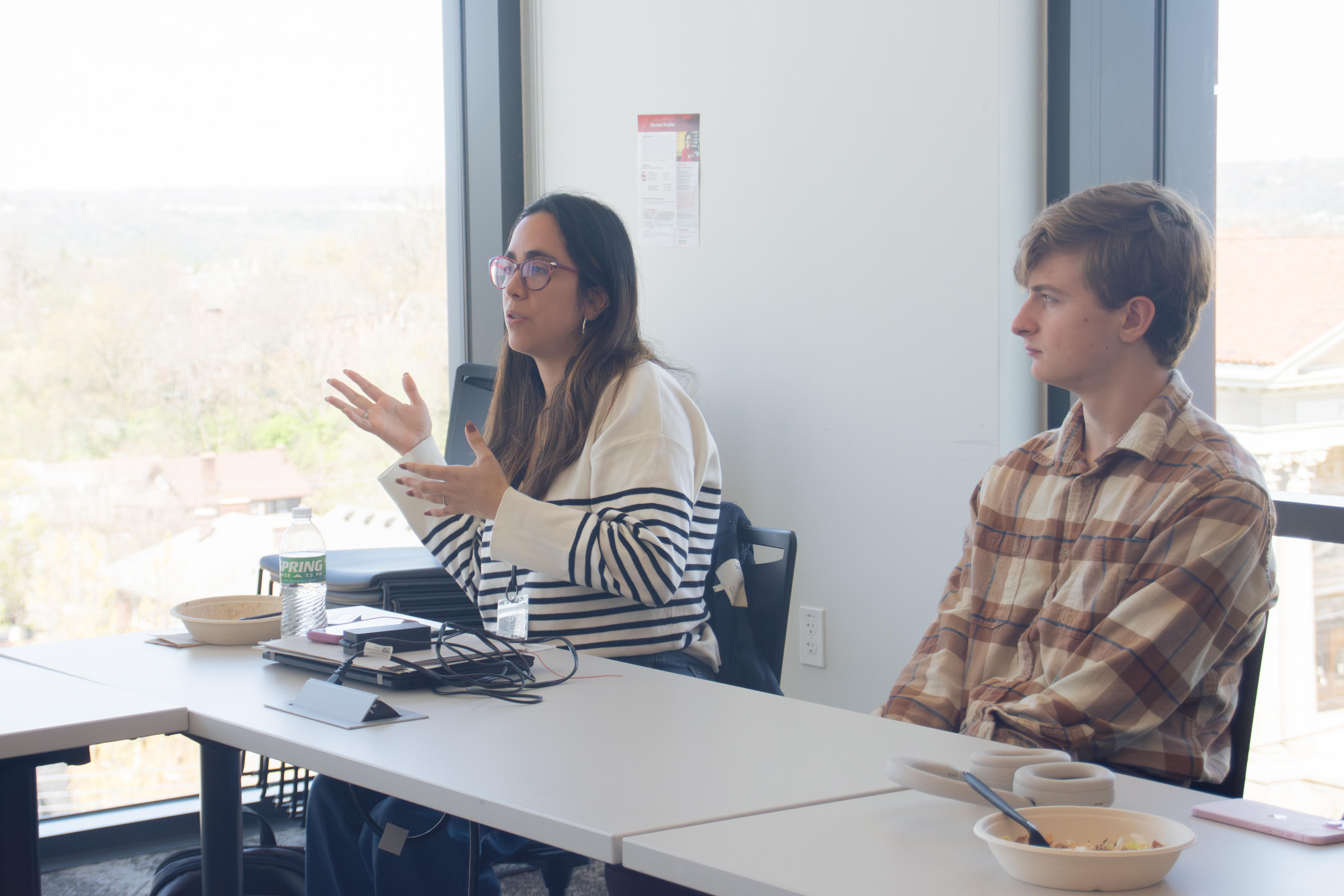 Two people are seated at a table in a bright room with large windows. One, wearing glasses and a striped shirt, is gesturing while speaking. The other listens attentively, wearing a plaid shirt. A water bottle, notebooks, and headphones are on the table.