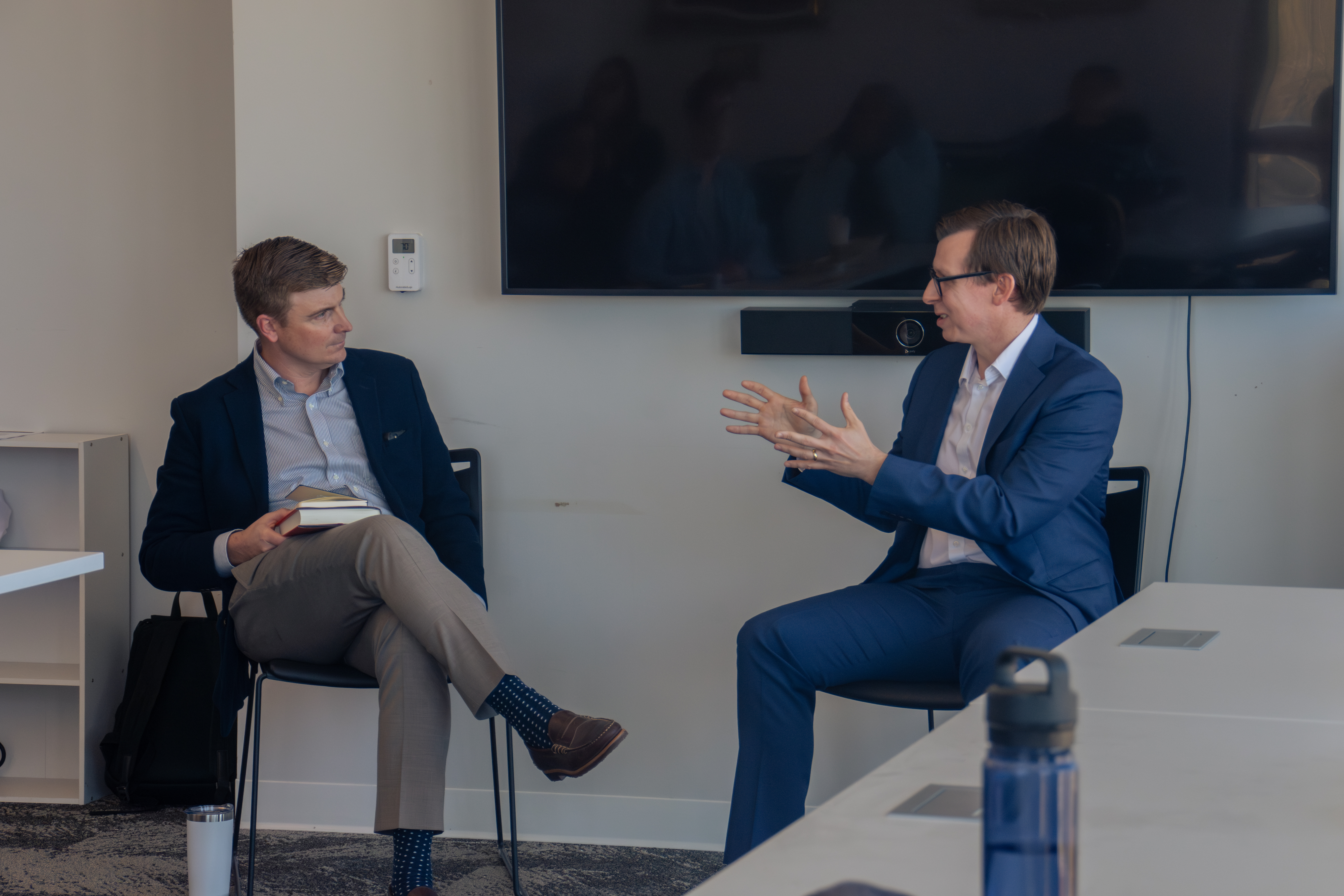 Two men in suits are seated in a modern office. One is gesturing passionately while speaking, as the other listens attentively, holding books.