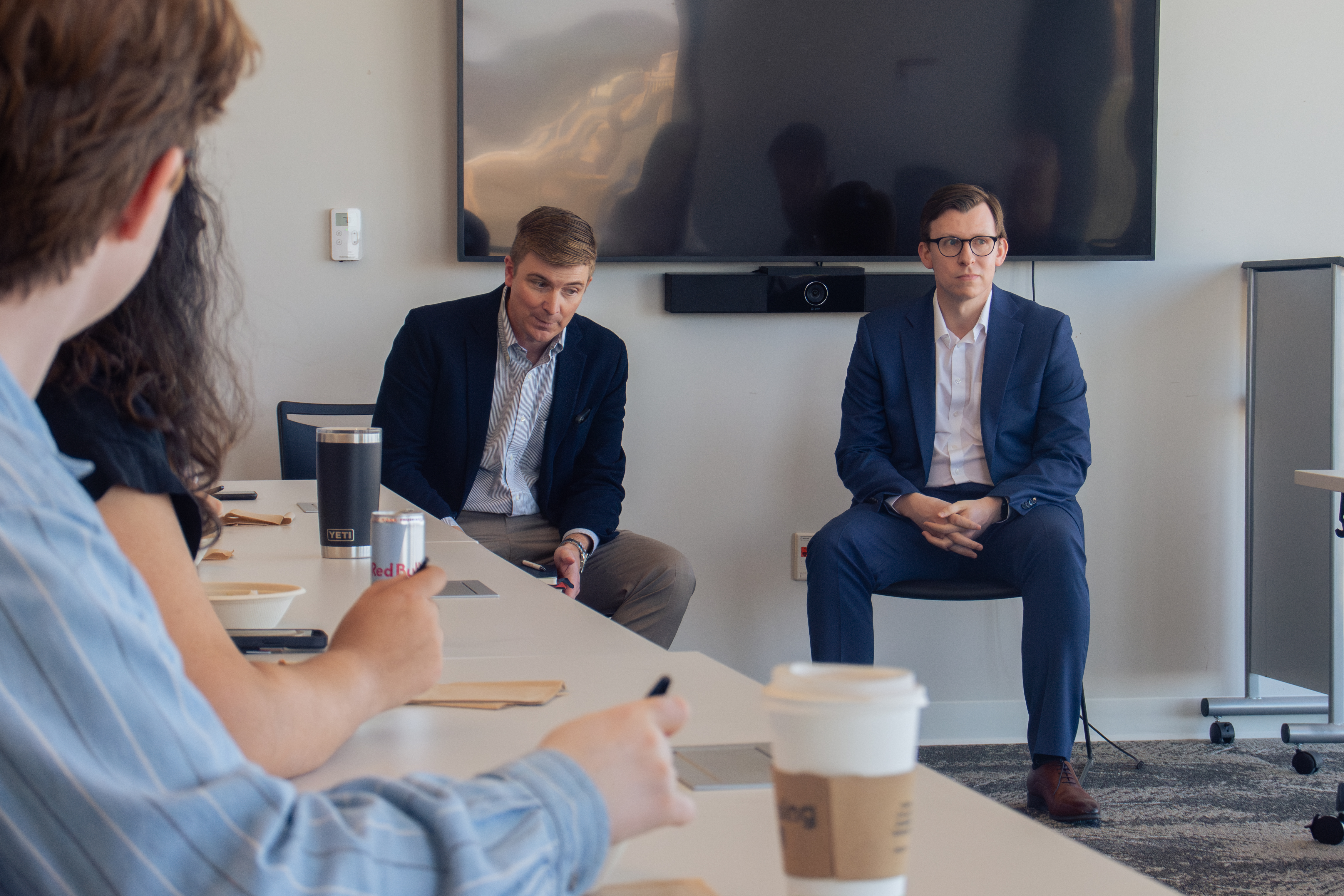 Two men in suits sit at the front of a conference room, addressing a group. Attendees hold coffee and energy drinks. The atmosphere is professional and focused.