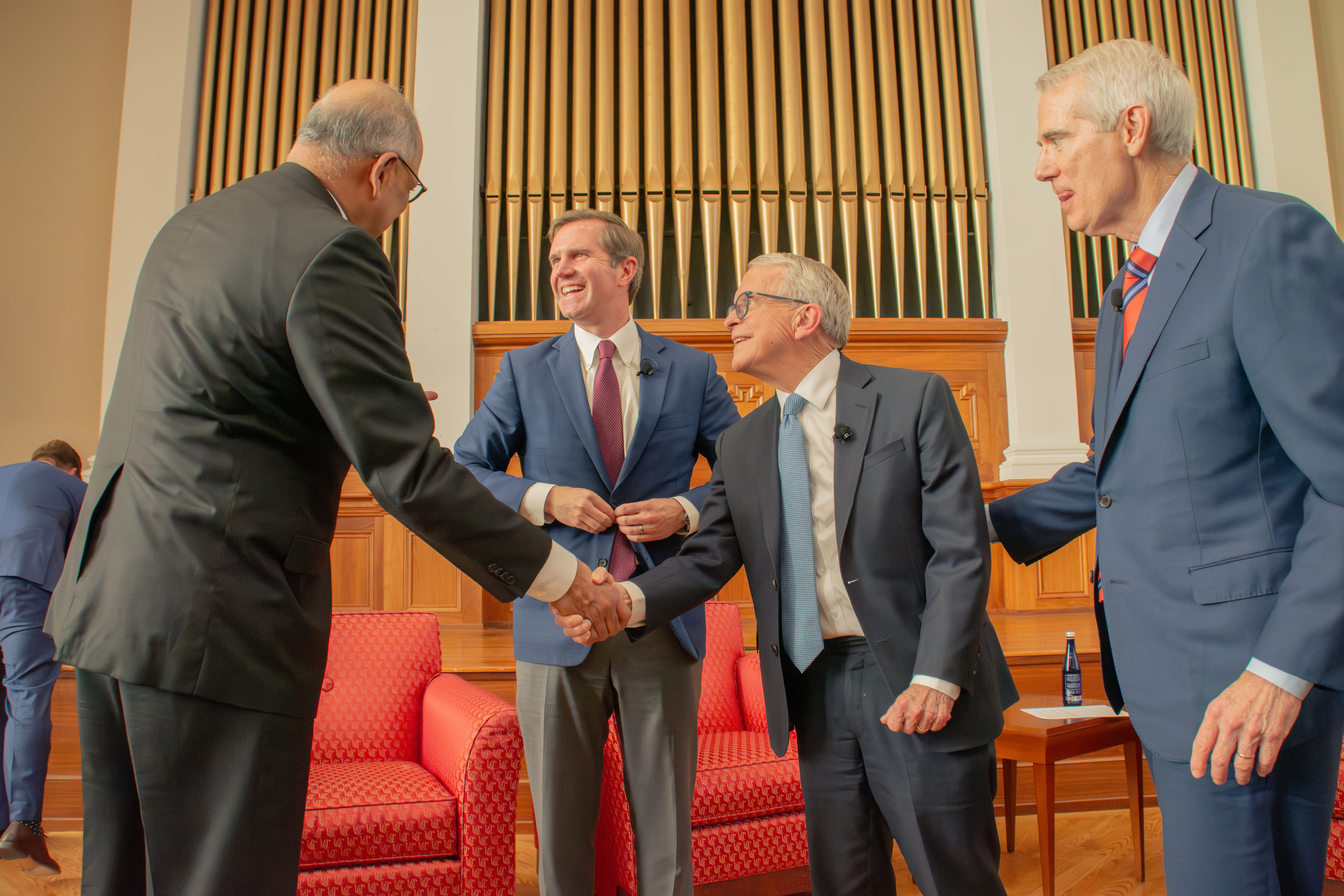 Four men in suits interact warmly in a formal setting, featuring red chairs and a wooden organ backdrop, conveying a sense of professionalism and camaraderie.