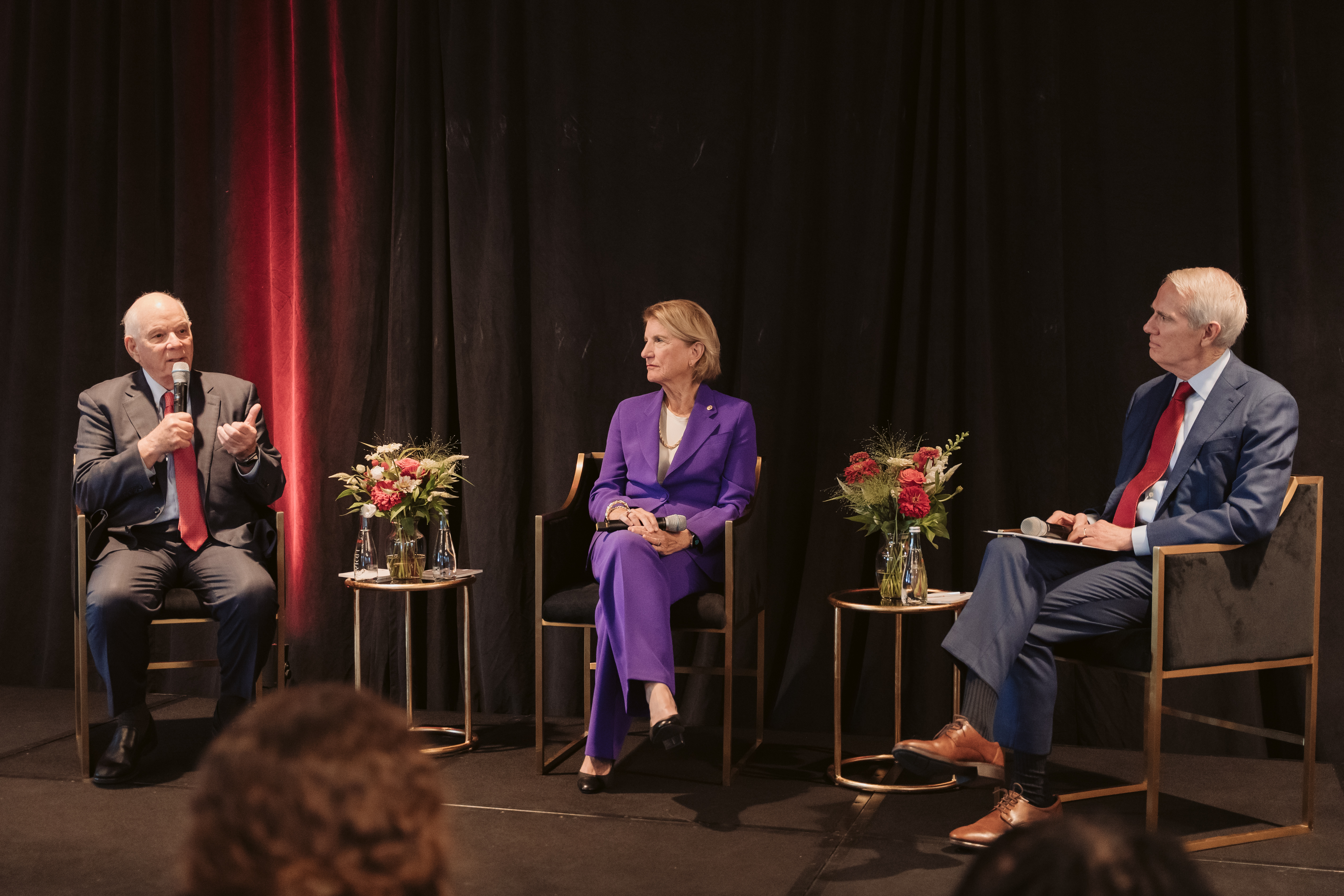 Three people, seated in chairs on stage with dark curtains, engage in discussion. They are in business attire, surrounded by small tables with flower arrangements, suggesting a formal, collaborative event.