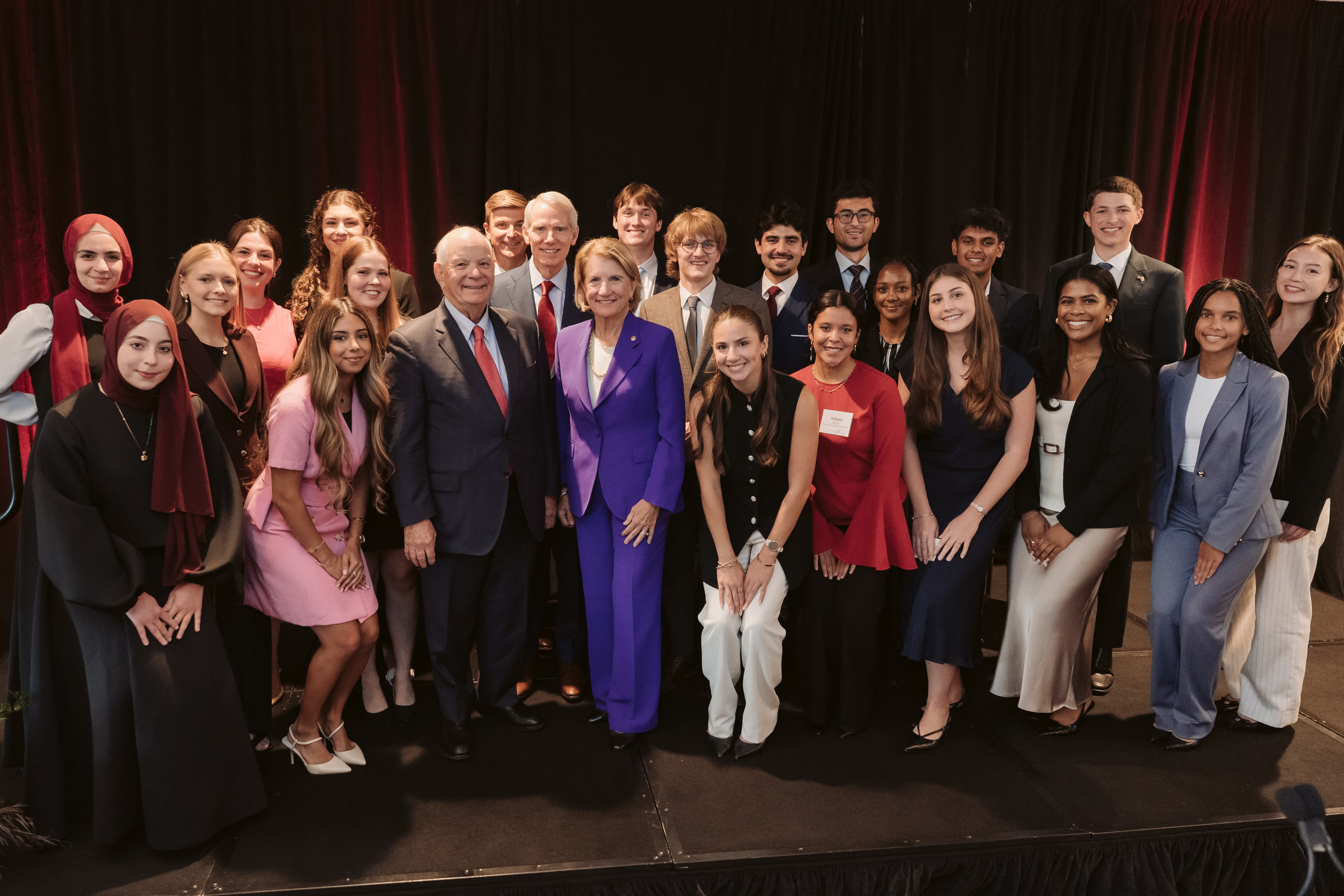 A diverse group of professionals, smiling and posing on stage, includes men and women in formal attire. The tone is celebratory and inclusive.