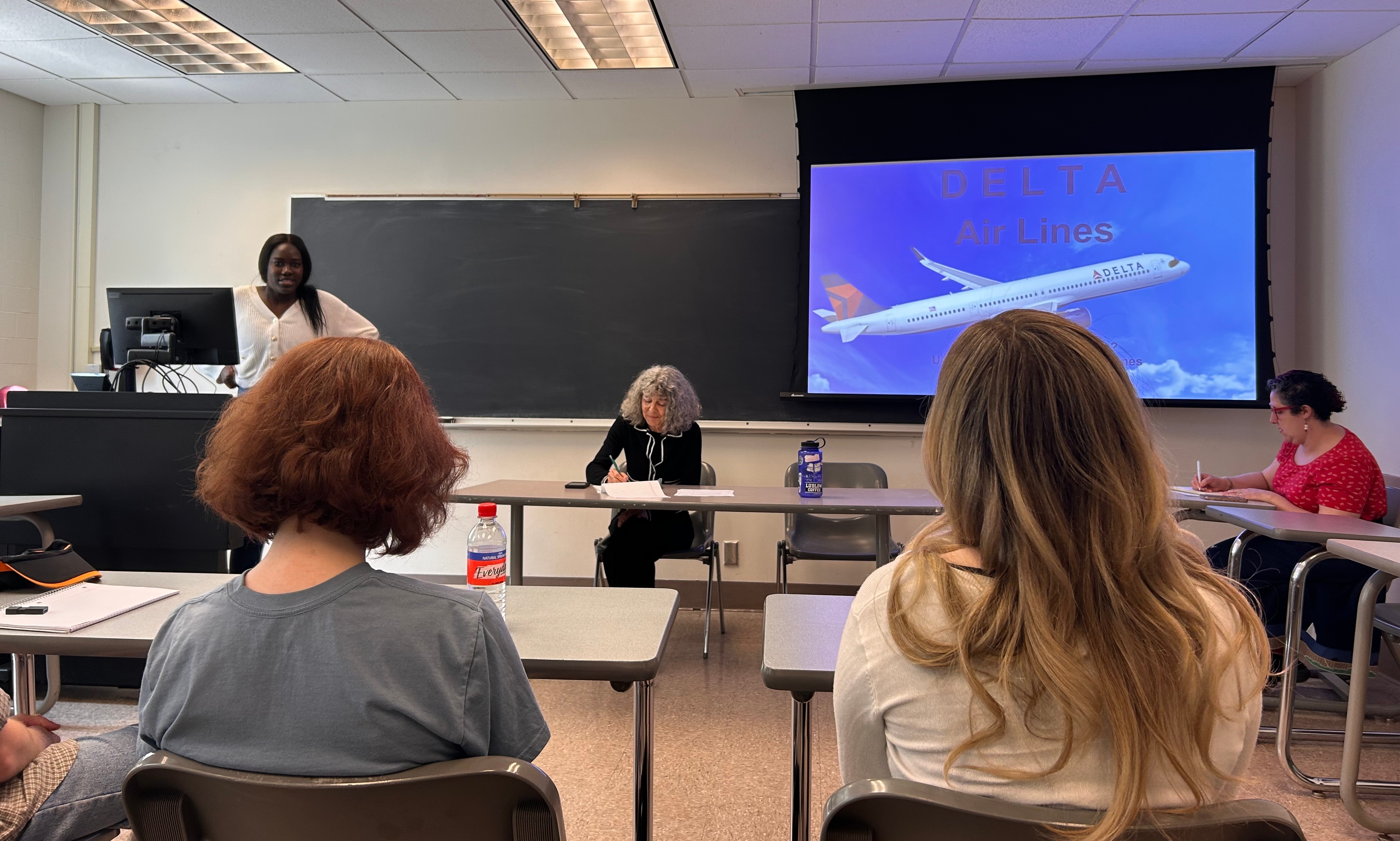 Students sitting in desks facing the front of a room. A blue screen is in the corner.