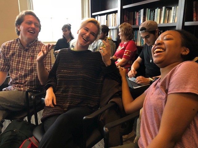 Three people laughing while seated in a room with books with more people in the background